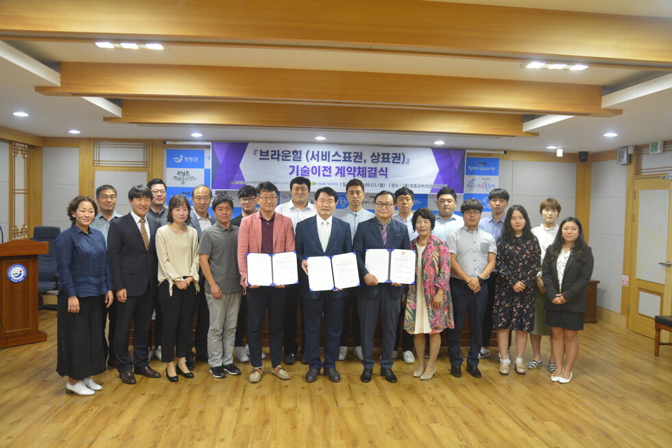 장흥군청 상황실에서 분홍색 자켓을 입은 남자와 양복을 입은 2명의 남자분이 상장을 들고 뒤로는 여러 사람들이 서서 건강기능식품 유통전문회사인 ㈜코리아바이오시스템와 연구개발 성과물의 상표권 및 서비스표권에 대한 이전계약을 체결을 기념하는 단체 사진