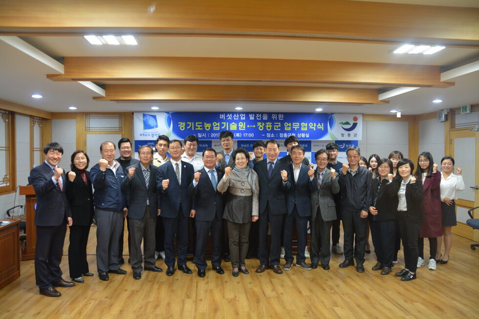 장흥군청 상황실에서 여러 양복을 입은 사람들이 한손에 주먹을 지며(화이팅 포즈를 하며) 경기도농업기술원(원장 김순재)과 버섯산업 발전을 위한 업무협약을 체결하는 기념 단체 사진