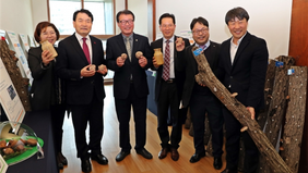 왼쪽에는 병을 든 여자분과 양손에 버섯 또는 병과 버섯을 들고 있는 남자 4명 그리고 버섯 재배 나무를 들고있는 남자분들이 버섯재배 기념 사진을 찍는 모습을 담은 사진