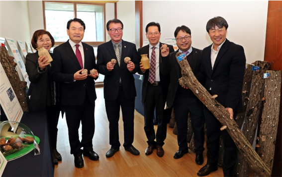 왼쪽에는 병을 든 여자분과 양손에 버섯 또는 병과 버섯을 들고 있는 남자 4명 그리고 버섯 재배 나무를 들고있는 남자분들이 버섯재배 기념 사진을 찍는 모습을 담은 사진