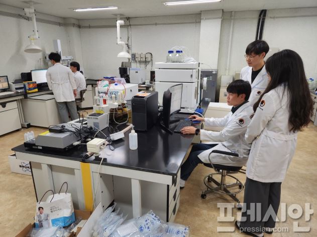연구실에서 하얀색 가운을 입고있는 남자 2명이 모니터를 보는 모습과 하얀색 가운을 입은 남자 1명이 컴퓨터를 만지고 뒤에는 하얀색 가운을 입은 남자 1명과 여자 1명이 그를 지켜보고 있는 모습을 담은 사진